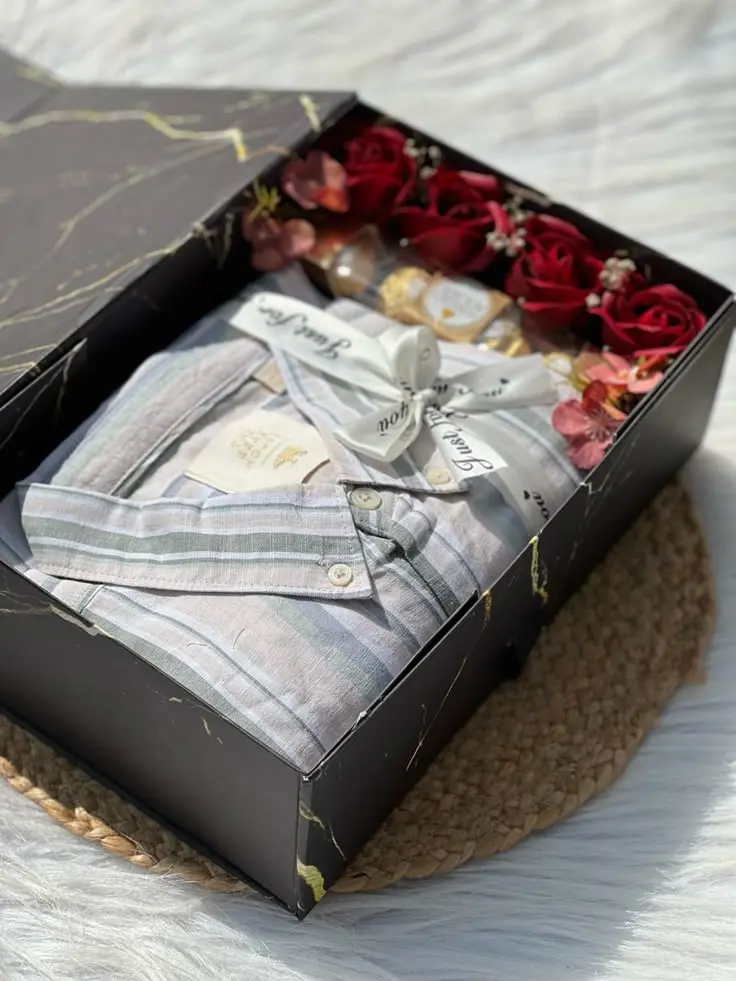 Customized Gifts & Services in Thrissur A neatly arranged gift hamper in a black box, featuring a folded striped shirt tied with a ribbon, red roses, small decorative flowers, and a perfume bottle.